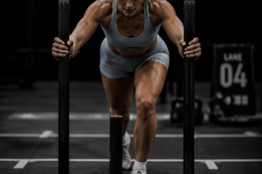 Blonde female athlete wearing a light blue sports bra and matching shorts pushing a weighted sled during a functional fitness competition in a dark, high-contrast indoor gym.
