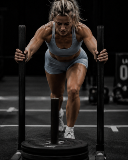 Blonde female athlete wearing a light blue sports bra and matching shorts pushing a weighted sled during a functional fitness competition in a dark, high-contrast indoor gym.