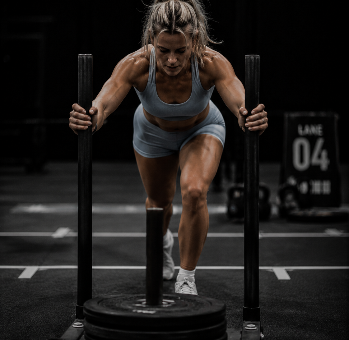 Blonde female athlete wearing a light blue sports bra and matching shorts pushing a weighted sled during a functional fitness competition in a dark, high-contrast indoor gym.